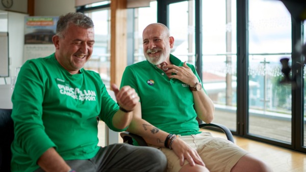 Two volunteers laughing together at the Macmillan Horizon Centre wearing Macmillan shirts. One man is holding a valve on his throat. 
