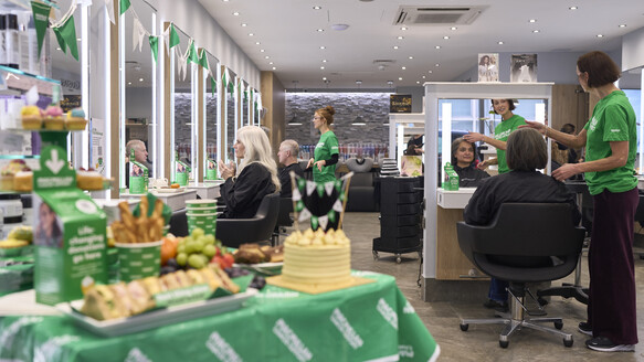 A Headmasters hairdressers is hosting a Coffee Morning. There is a table decorate for Coffee Morning. Hairdressers are wearing green Macmillan branded t-shirts and are at different stations across the salon talking to clients.