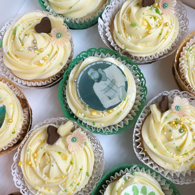 A close up of cupcakes. They are decorated with white frosting.  Some of them have edible images on them and other decorations like chocolate hearts, sprinkles and flowers.