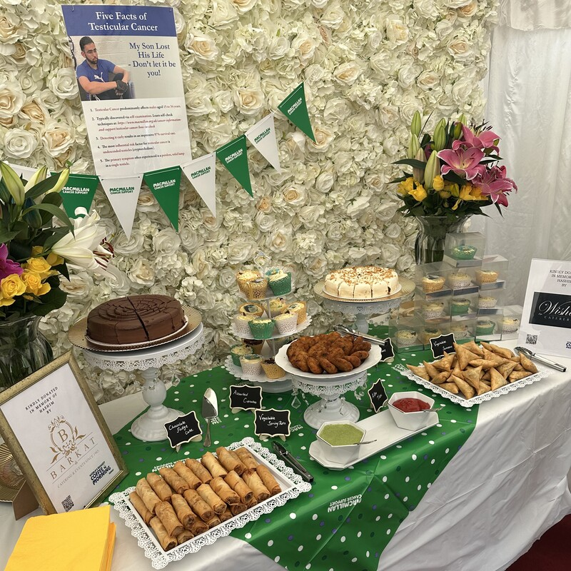 A table is decorated for a Coffee Morning. It has different sweet and savoury treats on it that are labelled. Behind the table is a white rose flower wall. On the wall is a poster about testicular cancer.