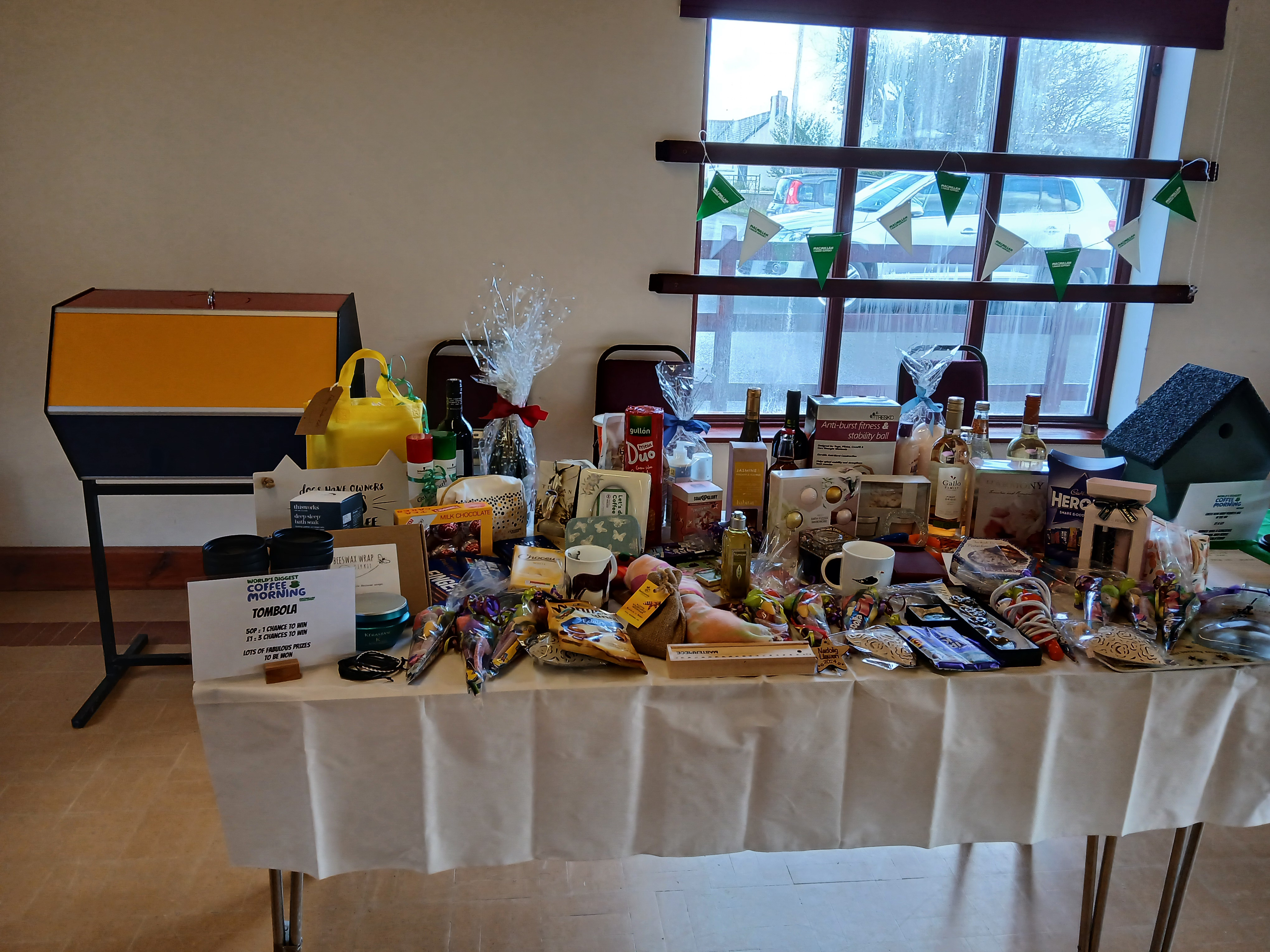 Many prizes are on a table inside a community hall. The prizes are for a tombola at a Coffee Morning.
