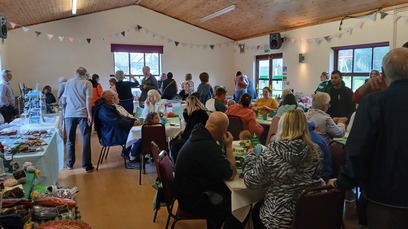 A village community hall is full of people taking part in a Coffee Morning. There are tables where people are sitting down and talking. There are other tables with food and raffle prizes.