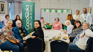 A group of people are smiling at the camera. Some are sat down in chairs at a table. Others are standing behind the table. They are indoors and have decorated for a Coffee Morning.