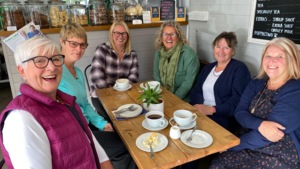 A group of people are sitting around a table in a café. They are smiling at the camera. Behind them is a display of different cakes and cookies.