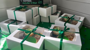 Several treat boxes are placed on a table, some stacked on one another. They have cupcakes and other treats inside them. There is also a sign with information about the sweet treat boxes with a price.