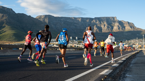 A group of runners taking part in a marathon. They are running along a road towards a settlement. In the distance is a hilly landscape. ©Mark Sampson