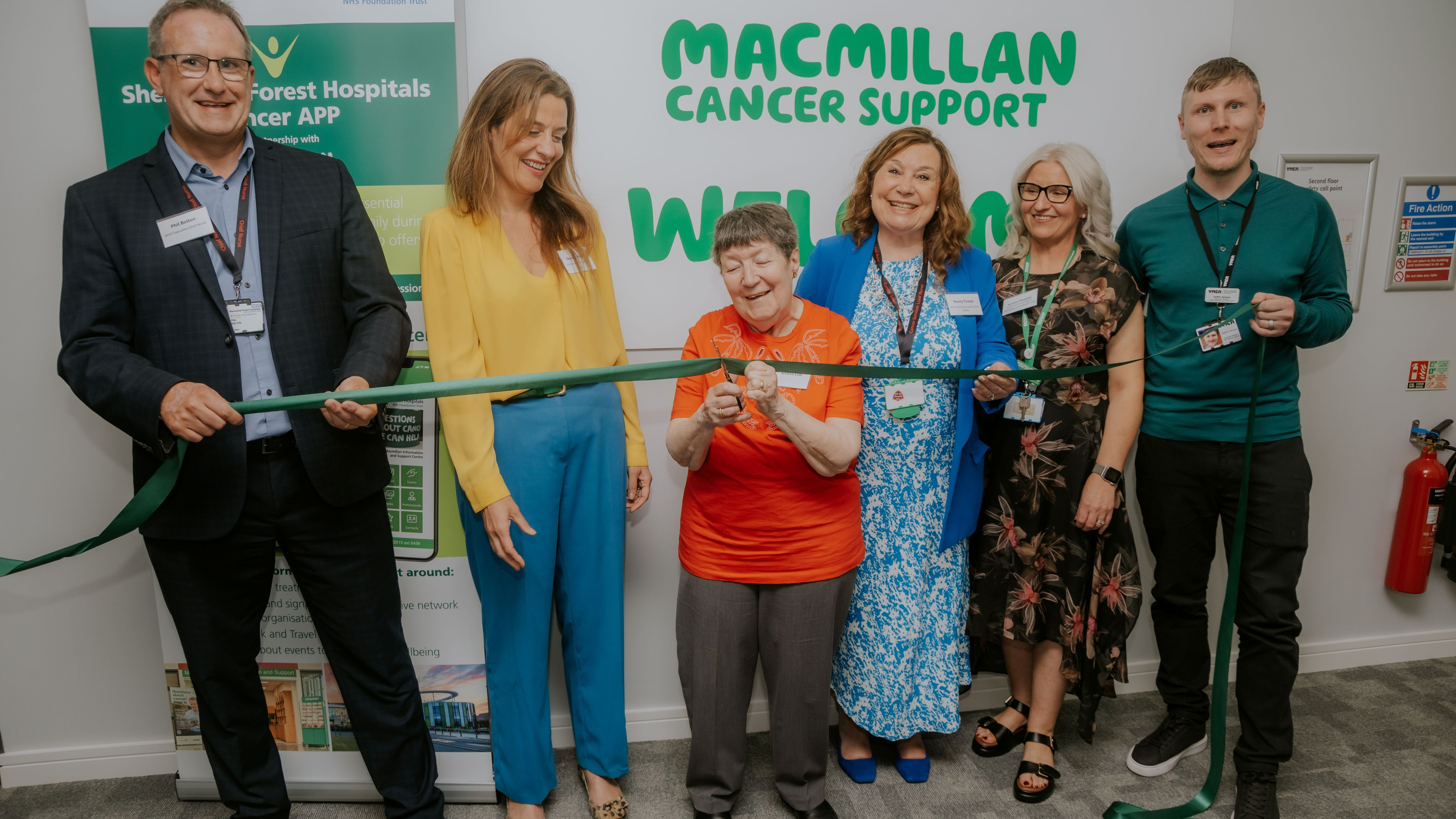 The photo is of six people holding a long green ribbon. The person in the middle is cutting the ribbon. The phots was taken at the opening of a community-based Macmillan Information & Support Service in Newark.