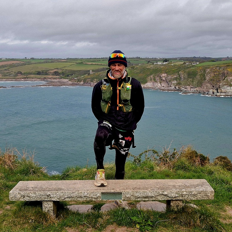 Ben has one leg perched on top of a stone bench. He is standing outside and behind him is a large body of water. He has on running gear including a running vest and trainers.