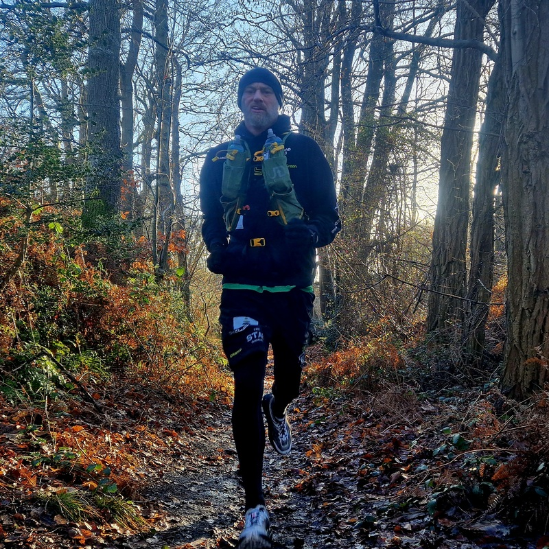 Ben is running along a path covered in fallen leaves. He is wearing a running vest, a long top and trousers, a hat and trainers.