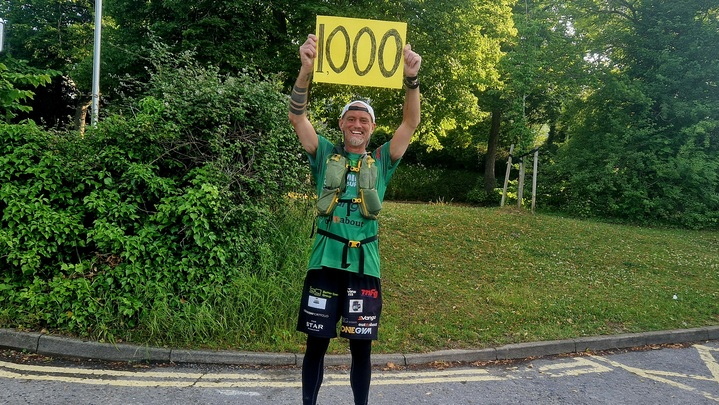 Ben is standing with his arms in the air holding a yellow sign that says 1000 on it. He is outside and is wearing different running gear.