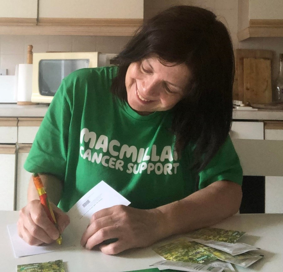 Woman signing Good Luck cards while smiling at a kitchen table and wearing a Macmillan t-shirt