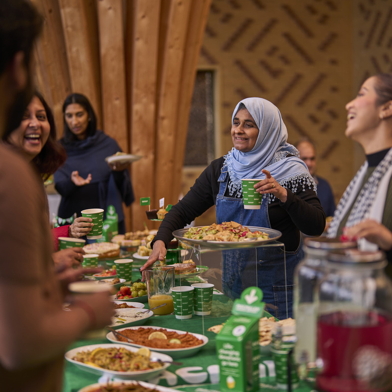 Many people are standing around a table decorated for a Coffee Morning. They appear to be talking to one another. One person in a blue hijab is holding a green Coffee Morning branded cup and pointing at some food on the table.