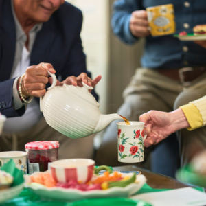 A person is tipping a white teapot to pour a warm drink into a floral mug that is being held by another person.