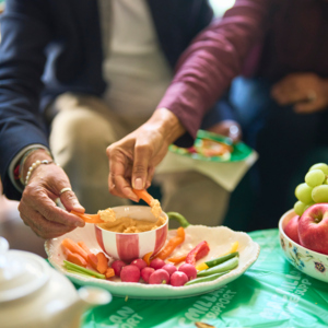 People are dipping carrots into hummus in a red and white stripped bowl. The bowl is placed on a large white plate that has different vegetables on it like radish and peppers. Next to the plate is a bowl with fruit in it like grapes and apples.