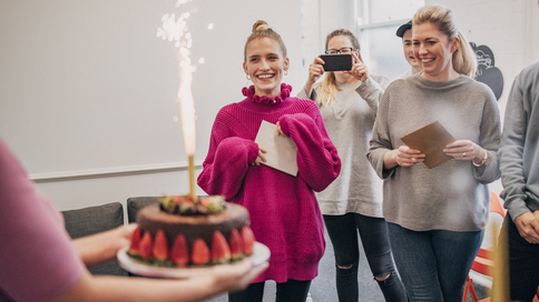 People are standing in an office and watching someone walk over holding a chocolate cake. The decadent cake is covered in strawberries. Many people are holding cards in their hands. One person is taking a photo with their phone. Photograph: SolStock/Getty Images