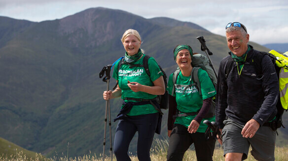 Three hikers are walking together. They are all carrying a rucksack on their back. Two of the hikers are wearing Macmillan branded tops. They appear to be hiking near a mountain.