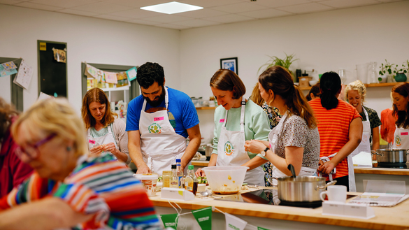 A group of people are gathered around a food preparation counter. There are different ingredients on the counter. The people are wearing white cooking aprons. 