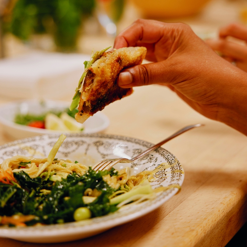 A person is holding a savoury pastry item in their hand. It is over a plate that has noodles and green veggies on it. Photography: Rebecca Naen