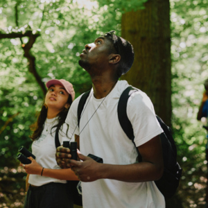 Two people are standing outside in the woods. Both are holding binoculars. One person is looking up to the trees. Photography by: Rebecca Naen.
