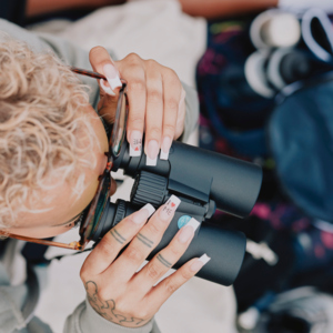 A view from above of a person holding a pair of binoculars. They have short, blonde curly hair. They are wearing a light grey jacket and have their nails done. Photography by Rebecca Naen.