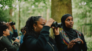 A group of people are bird watching in the woods. They are holding binoculars. Photography by Rebecca Naen.