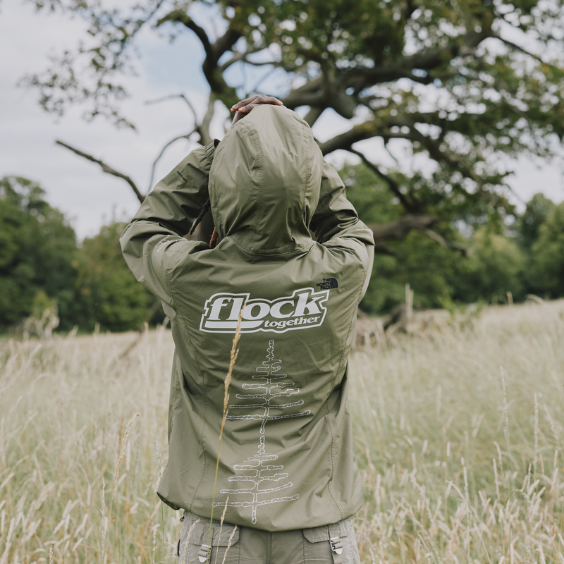 A person is standing in a field. They are wearing a light green jacket that has the logo Flock Together on it. They appear to be looking up towards a tree in the field. Photography by: Rebecca Naen.