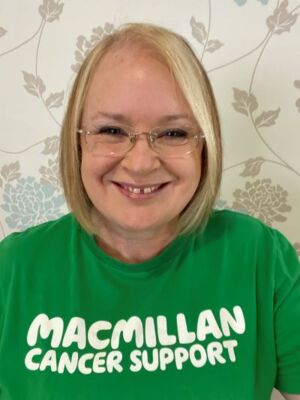 Carrol is smiling at the camera, she has blonde hair and is wearing glasses. She is wearing a green t-shirt which reads Macmillan Cancer Support.