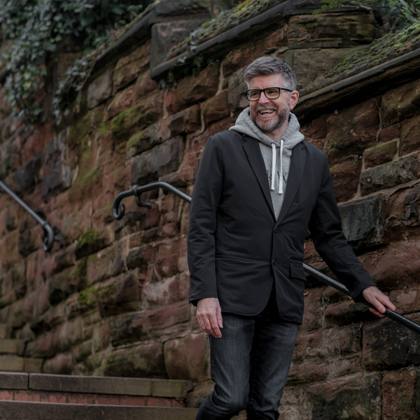 Phil is walking down a stair case alongside a brick wall. He has short hair and is wearing black frammed glasses, a dark jacket, grey hoodie and dark trousers. 