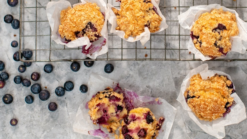 Golden-brown blueberry muffins with a crumb topping rest on a cooling rack, wrapped in white parchment paper. Scattered blueberries and visible moist interiors make these treats look irresistibly fresh and delicious.