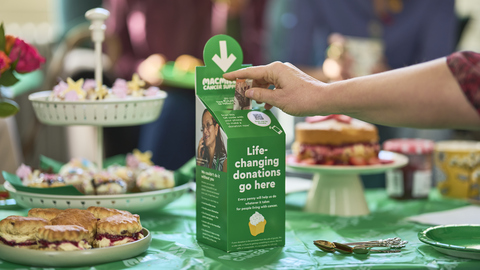 A person is donating £10 in a Coffee Morning donation box. It is on a table full of delicious bites and treats.