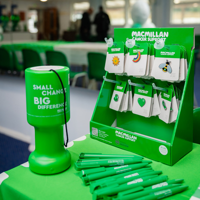 A display with several Macmillan pins on them. In front is a green charity box and many Macmillan branded pens.
