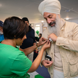 Pam is pinning a Macmillan badge on another person's top. Pam is wearing a bright green top and is on the left. The person who is receiving the badge is wearing a tan coloured top and jacket. They are also wearing a tan dastar/dumalla.