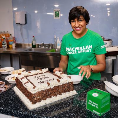 Pam is standing behind a marble topped counter that has a large cake on it. Pam has short dark hair and is wearing a Macmillan branded green top. The cake has been decorated with white and brown frosting and has the words 'Pam's Coffee Morning' on them.