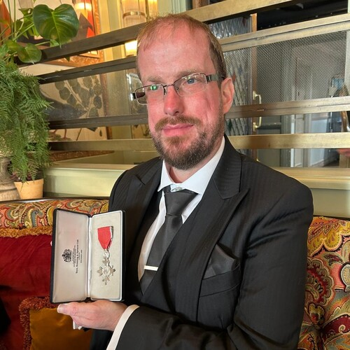 Nathaniel is holding his MBE. He has short hair and is wearing a dark suit. 