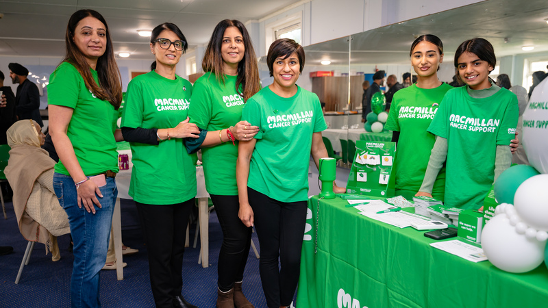 Pam is standing in the middle of a group of other people who are volunteering at her Coffee Morning. They are all wearing a green Macmillan branded. Some are stood behind a table that has different Coffee Morning donate asks and cancer information.