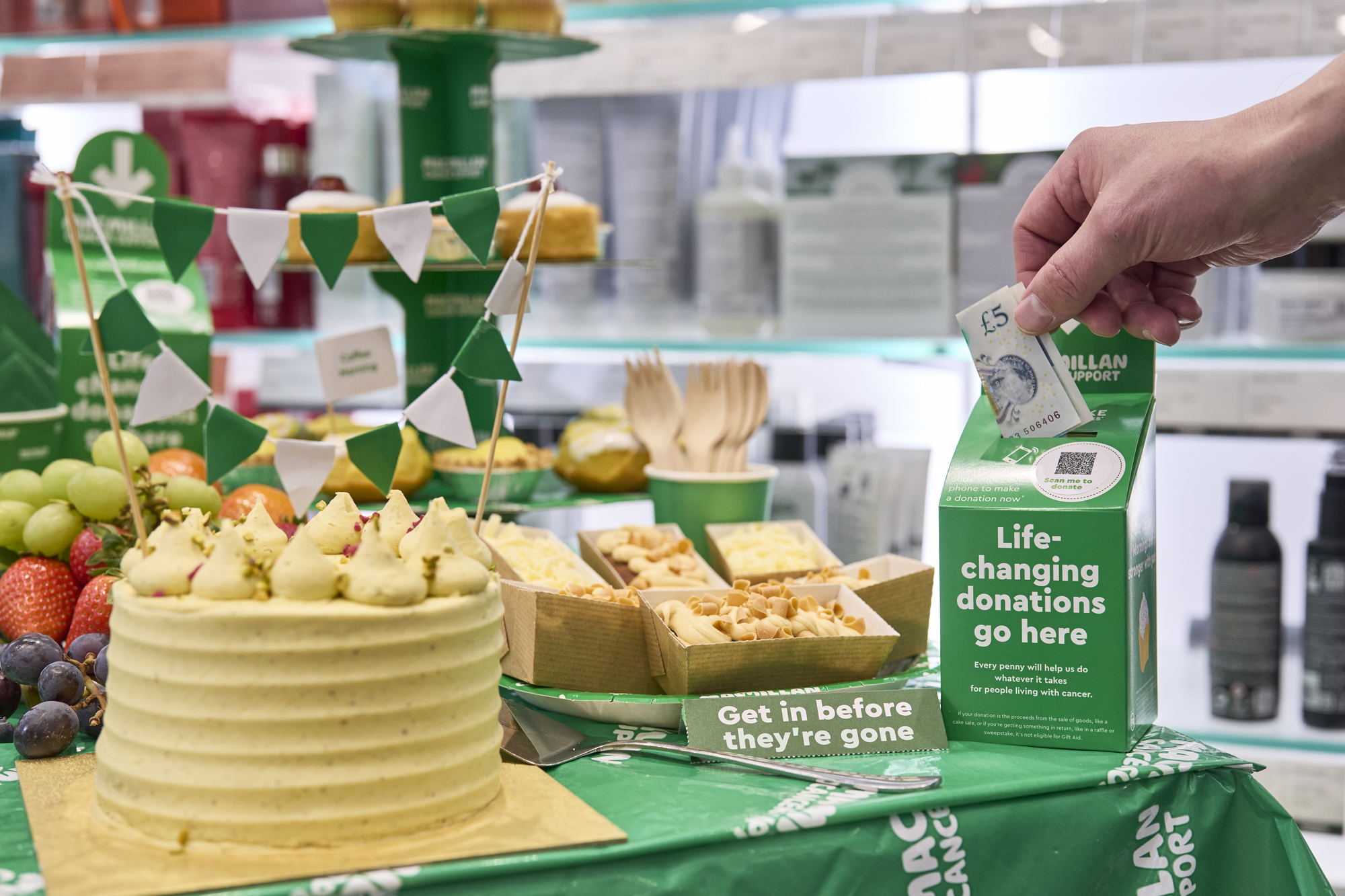 A Coffee Morning table with cakes on and a hand putting money into a donation box