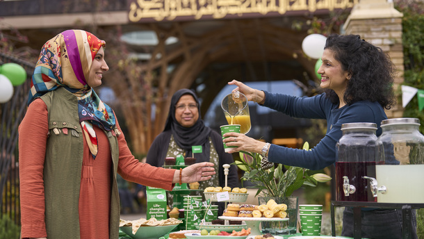 A person is pouring juice into a cup to give to someone. They are outside a mosque and are celebrating Coffee Morning. There are different treats on the table in front of them that has been decorated for the event.