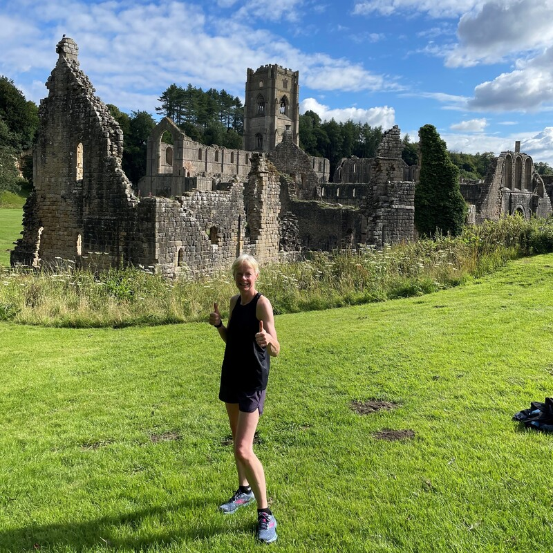Vicky is giving two thumbs up as she stands in front of some castle ruins. She is wearing a running vest top and shorts. 
