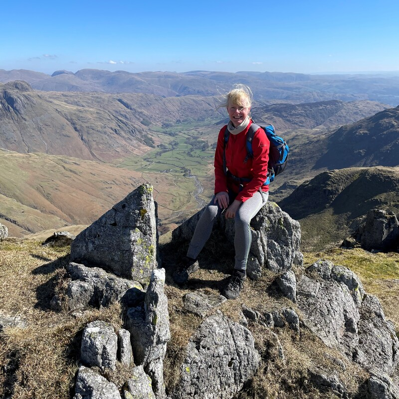 Vicky is sitting down on a boulder on top of a mountain. She is wearing a bright coat, long trousers and a rucksack. Behind her is a view of a valley.