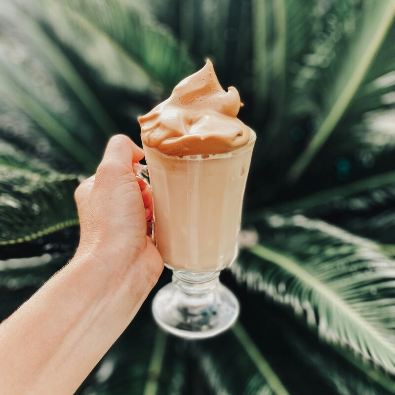 A person is holding up a clear latte glasses full to the brim with dalgona coffee. It has a whipped stiff peak on top that is overflowing. Photo by Chelsea Audibert on Unsplash.