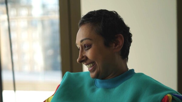 A woman with a colourful top, short hair and bright eye make-up is smiling