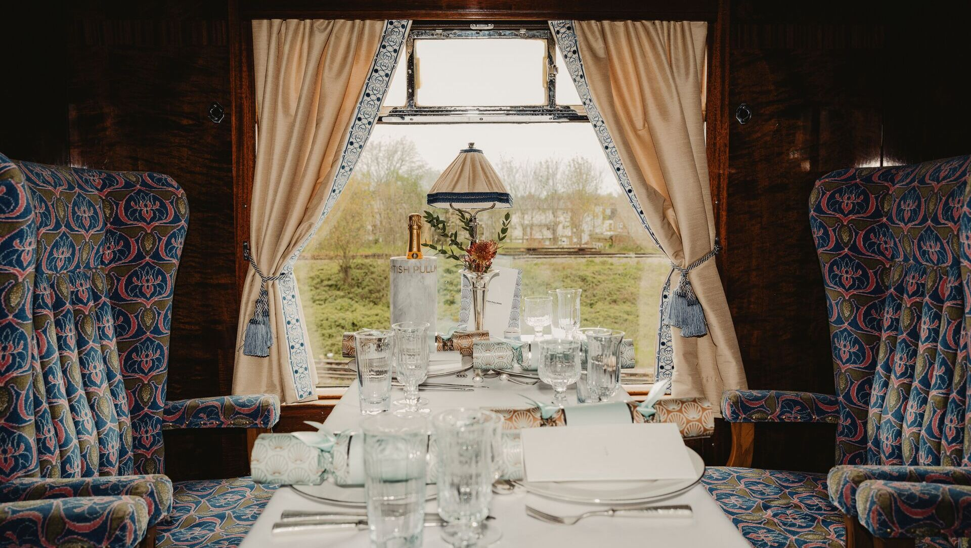 A view of the inside of the Belmond British Pullman. There are two comfortable armchairs across from another with a table in between. There is a window next to the chairs showing the countryside as the trains moves. Tables includes a white table cloth, place settings and a Champaign cooler with a bottle in it. 