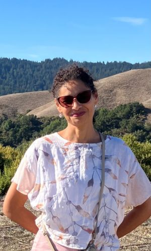 Woman with sunglasses standing outdoors. In the background there are trees and hills. Sher is wearing a white top with print.