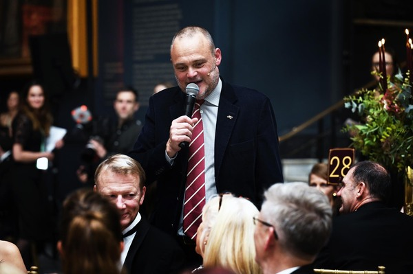 Comedian Al Murray is speaking into a microphone at a Macmillan Ball. He is standing near a group of people sitting down. Al is wearing a dark suit with a red and white stripped tie. He has short hair and a beard.