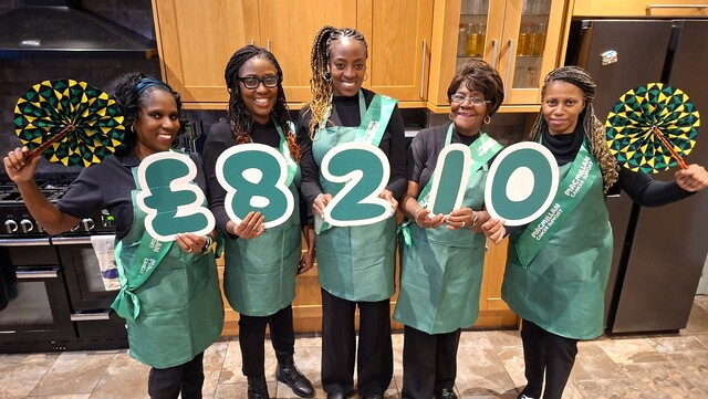 Marcia is standing in a row with other members of her family. They are all wearing green aprons and black tops and trousers. Each person is holding up a cardboard cut out with the amount that has been raised. They are standing in a kitchen.