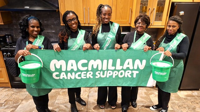 Marcia is standing in a row with her family. They are holding up a Macmillan Cancer Support banner. They are wearing black tops and trousers, and a green apron and sashes. They are standing in a kitchen.
