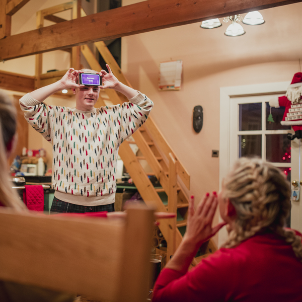A group of people are celebrating the Christmas period with games. One person is standing up and has their mobile phone on their forehead. The screen is facing others at the party who are giving clues to the person holding the phone what the screen may say.