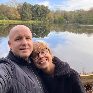 Rachel is a young woman who was diagnosed with bowel cancer. She is smiling and taking a selfie with her partner in front of a lake surrounded by trees.