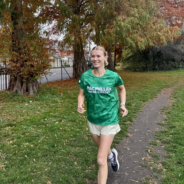 A person running along a paved path in a park. They have short blonde hair that is tied up and are wearing a green Macmillan branded running top. 