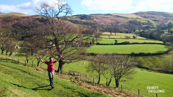 A hiker standing in a grassy field with both arms in the air. Behind him are grassy fields and rolling hills.